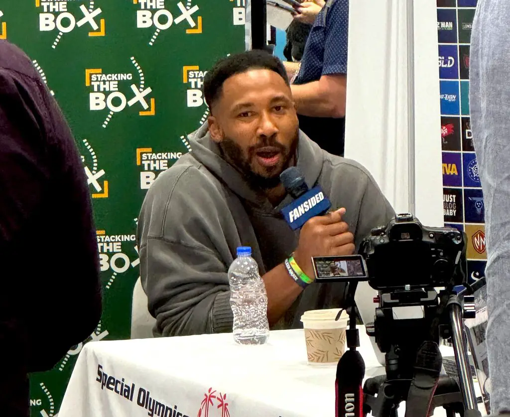 Mayles Garrett in einem grauen Kapuzenpulli spricht in ein Mikrofon mit der Aufschrift „FANSIDED“. Sie sitzen mit einer Wasserflasche und einer Kaffeetasse an einem Tisch und unterhalten sich über Myles Garrett. Dahinter ist ein grün-weißer „STACKING THE BOX“-Hintergrund mit Logos zu sehen, und im Vordergrund fängt die Kamera den Geist Detroits ein. Diese Beschreibung wurde mit der FootballR KI automatisch generiert.