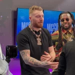 Drei Personen unterhalten sich in einem Innenraum neben einem weißen Fahrzeug, das voller Unterschriften ist. Die Person links, die mit ihren Tattoos und einer großen Halskette Maxx Crosby ähnelt, gestikuliert beim Sprechen. Die Person in der Mitte trägt eine Sonnenbrille und ein gestreiftes Hemd. Eine Person mit kurzen Haaren blickt weg. Im Hintergrund sind zwei Bildschirme zu sehen. Diese Beschreibung wurde mit der FootballR KI automatisch generiert.