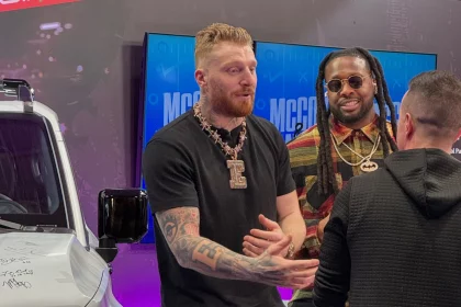 Drei Personen unterhalten sich in einem Innenraum neben einem weißen Fahrzeug, das voller Unterschriften ist. Die Person links, die mit ihren Tattoos und einer großen Halskette Maxx Crosby ähnelt, gestikuliert beim Sprechen. Die Person in der Mitte trägt eine Sonnenbrille und ein gestreiftes Hemd. Eine Person mit kurzen Haaren blickt weg. Im Hintergrund sind zwei Bildschirme zu sehen. Diese Beschreibung wurde mit der FootballR KI automatisch generiert.