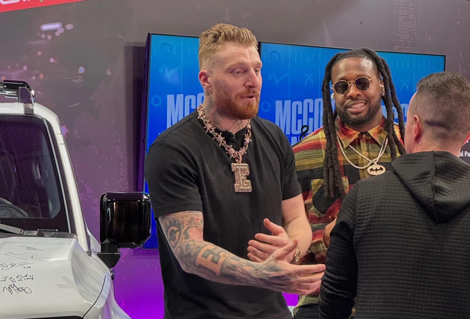 Drei Personen unterhalten sich in einem Innenraum neben einem weißen Fahrzeug, das voller Unterschriften ist. Die Person links, die mit ihren Tattoos und einer großen Halskette Maxx Crosby ähnelt, gestikuliert beim Sprechen. Die Person in der Mitte trägt eine Sonnenbrille und ein gestreiftes Hemd. Eine Person mit kurzen Haaren blickt weg. Im Hintergrund sind zwei Bildschirme zu sehen. Diese Beschreibung wurde mit der FootballR KI automatisch generiert.