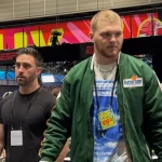 Aidan Hutchinson schlendert durch den NFL Media Radio Row Innenraum. Der rechte trägt eine grüne Jacke, ein blaues T-Shirt und einen VIP-Pass. Sein Begleiter links trägt ein schwarzes T-Shirt. ESPN-Banner und eine große Säule rahmen sie ein, während Myles Garrett im Hintergrund zwischen den Tischen und anderen Gästen verkehrt. Diese Beschreibung wurde mit der FootballR KI automatisch generiert.
