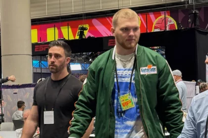 Aidan Hutchinson schlendert durch den NFL Media Radio Row Innenraum. Der rechte trägt eine grüne Jacke, ein blaues T-Shirt und einen VIP-Pass. Sein Begleiter links trägt ein schwarzes T-Shirt. ESPN-Banner und eine große Säule rahmen sie ein, während Myles Garrett im Hintergrund zwischen den Tischen und anderen Gästen verkehrt. Diese Beschreibung wurde mit der FootballR KI automatisch generiert.
