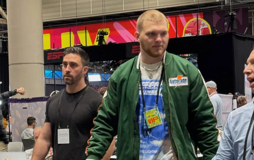 Aidan Hutchinson schlendert durch den NFL Media Radio Row Innenraum. Der rechte trägt eine grüne Jacke, ein blaues T-Shirt und einen VIP-Pass. Sein Begleiter links trägt ein schwarzes T-Shirt. ESPN-Banner und eine große Säule rahmen sie ein, während Myles Garrett im Hintergrund zwischen den Tischen und anderen Gästen verkehrt. Diese Beschreibung wurde mit der FootballR KI automatisch generiert.