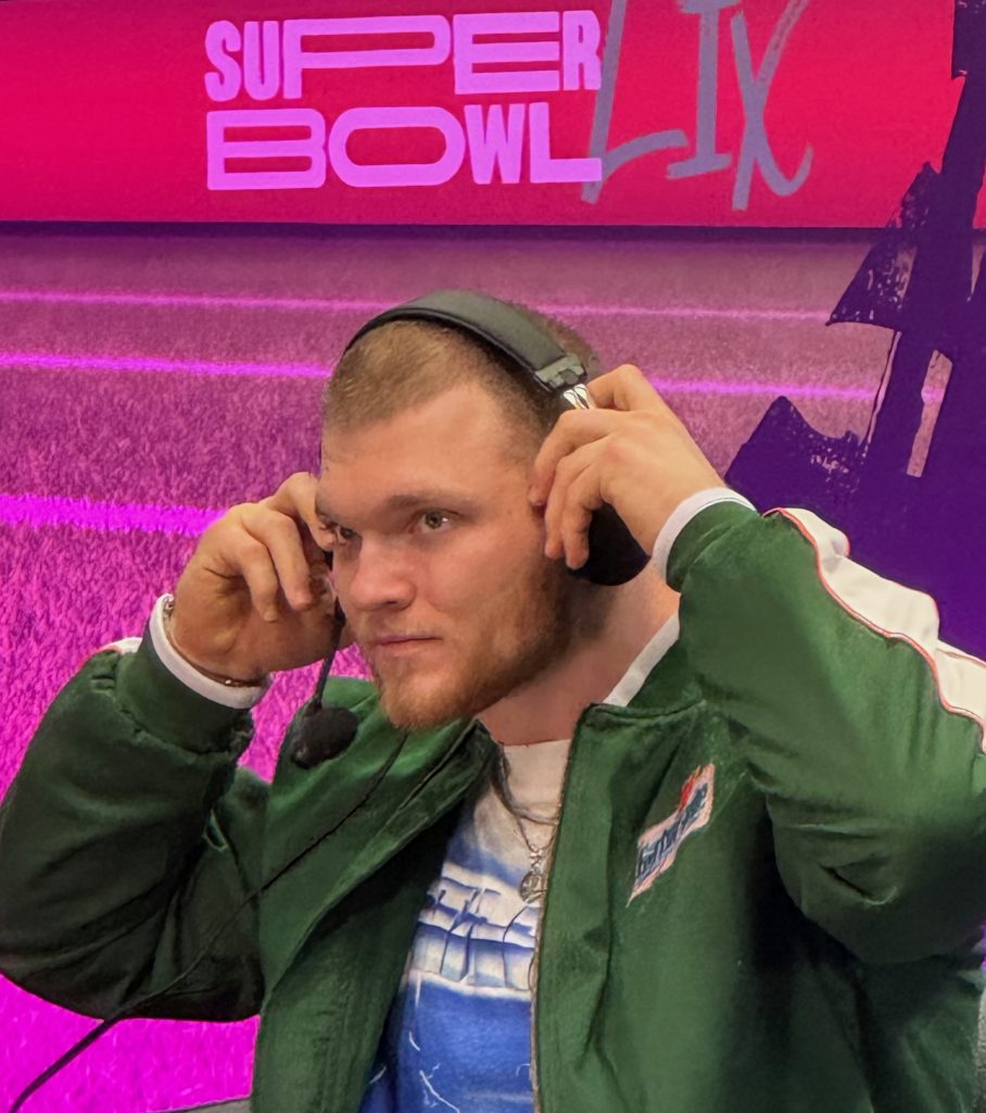 Aidan Hutchinson mit kurzen Haaren und Bart, die an Aidan Hutchinson erinnert, trägt Kopfhörer und Mikrofon und passt das Headset im Sitzen an. Sie trägt eine grüne Jacke über einem blauen Hemd vor einem violetten Hintergrund mit dem Text „SUPER BOWL LIX“, was auf Detroits lebhaften Footballgeist hinweist. Diese Beschreibung wurde mit der FootballR KI automatisch generiert.