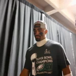 Super Bowl MVP Jalen Hurts mit verkehrt herum aufgesetzter Kappe und einem Philadelphia Eagles-T-Shirt mit der Aufschrift „Super Bowl Champions“ geht neben einem anderen Mann durch einen Flur. Schwarze Vorhänge säumen den Flur, und beide wirken konzentriert. Der zweite Mann, der ein orangefarbenes Abzeichen an einem Schlüsselband trägt, scheint ebenso konzentriert zu sein. Diese Beschreibung wurde mit der FootballR KI automatisch generiert.