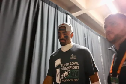 Super Bowl MVP Jalen Hurts mit verkehrt herum aufgesetzter Kappe und einem Philadelphia Eagles-T-Shirt mit der Aufschrift „Super Bowl Champions“ geht neben einem anderen Mann durch einen Flur. Schwarze Vorhänge säumen den Flur, und beide wirken konzentriert. Der zweite Mann, der ein orangefarbenes Abzeichen an einem Schlüsselband trägt, scheint ebenso konzentriert zu sein. Diese Beschreibung wurde mit der FootballR KI automatisch generiert.
