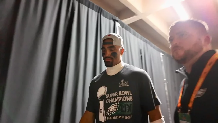 Super Bowl MVP Jalen Hurts mit verkehrt herum aufgesetzter Kappe und einem Philadelphia Eagles-T-Shirt mit der Aufschrift „Super Bowl Champions“ geht neben einem anderen Mann durch einen Flur. Schwarze Vorhänge säumen den Flur, und beide wirken konzentriert. Der zweite Mann, der ein orangefarbenes Abzeichen an einem Schlüsselband trägt, scheint ebenso konzentriert zu sein. Diese Beschreibung wurde mit der FootballR KI automatisch generiert.