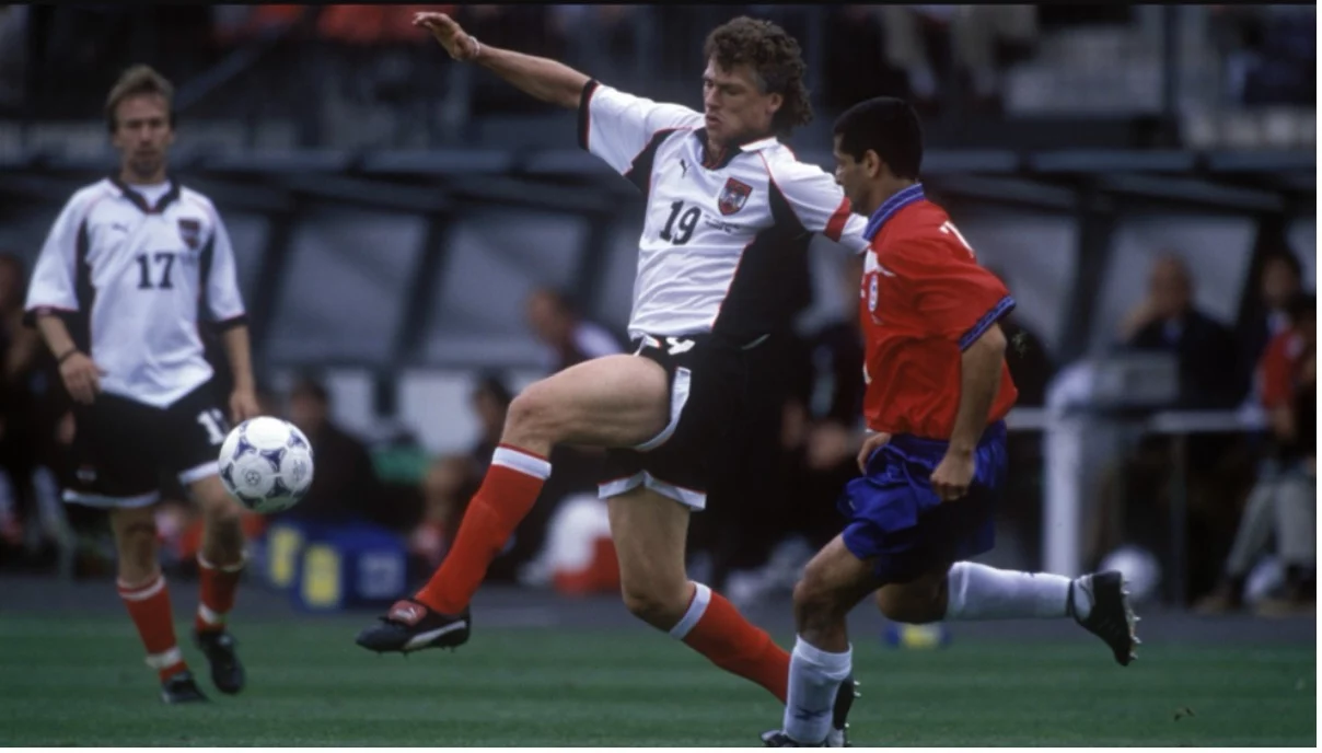 Zwei Fußballspieler in Aktion auf dem Feld. Der Spieler im weiß-schwarzen Trikot mit der Nummer 19 streckt sein Bein aus, um den Ball zu kontrollieren, was an einen Topstürmer wie Toni Polster erinnert. Ein gegnerischer Spieler im rot-blauen Trikot mit der Nummer 18 fordert ihn heraus, während ein weiterer im Hintergrund zu sehen ist. Diese Beschreibung wurde mit der FootballR KI automatisch generiert.