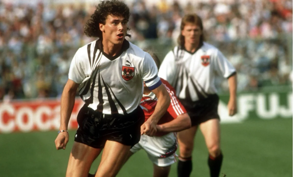 Ein Fußballspieler in einem weißen Trikot mit schwarzen Streifen läuft in schwarzen Shorts über das Feld. Er hat lockiges Haar und eine Kapitänsbinde, die an die österreichische Fußball-Ikone Toni Polster erinnert. Im Hintergrund ist ein weiterer Spieler mit langen Haaren zu sehen; eine Menschenmenge und rote Coca-Cola-Schilder sind zu sehen. Diese Beschreibung wurde mit der FootballR KI automatisch generiert.