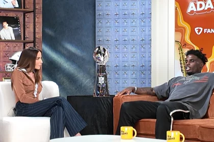 NEW ORLEANS - Media Row - Kay Adams und Tyreek Hill sitzen in einem Studio mit einer Sportkulisse mit Tyreek Hill. Die Frau trägt eine braune Jacke und gestreifte Hosen, während der Mann ein graues T-Shirt und schwarze Hosen trägt. Zwischen ihnen steht eine Trophäe. Auf dem Tisch davor stehen leuchtend orangefarbene Tassen mit einem Logo. Diese Beschreibung wurde mit der FootballR KI automatisch generiert.