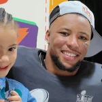 Ein lächelnder Saquon Barkley mit Bart, einer verkehrt herum aufgesetzten Kappe und einem dunklen Hemd mit dem Logo der Philadelphia Eagles posiert mit einem kleinen Mädchen. Das Mädchen mit geflochtenen Haaren und einem blauen Jeanshemd hält einen kleinen Gegenstand in der Hand. Sie sind auf einer Pressekonferenz mit einem farbenfrohen Hintergrund und einem NFL-Mikrofon. Diese Beschreibung wurde mit der FootballR KI automatisch generiert.