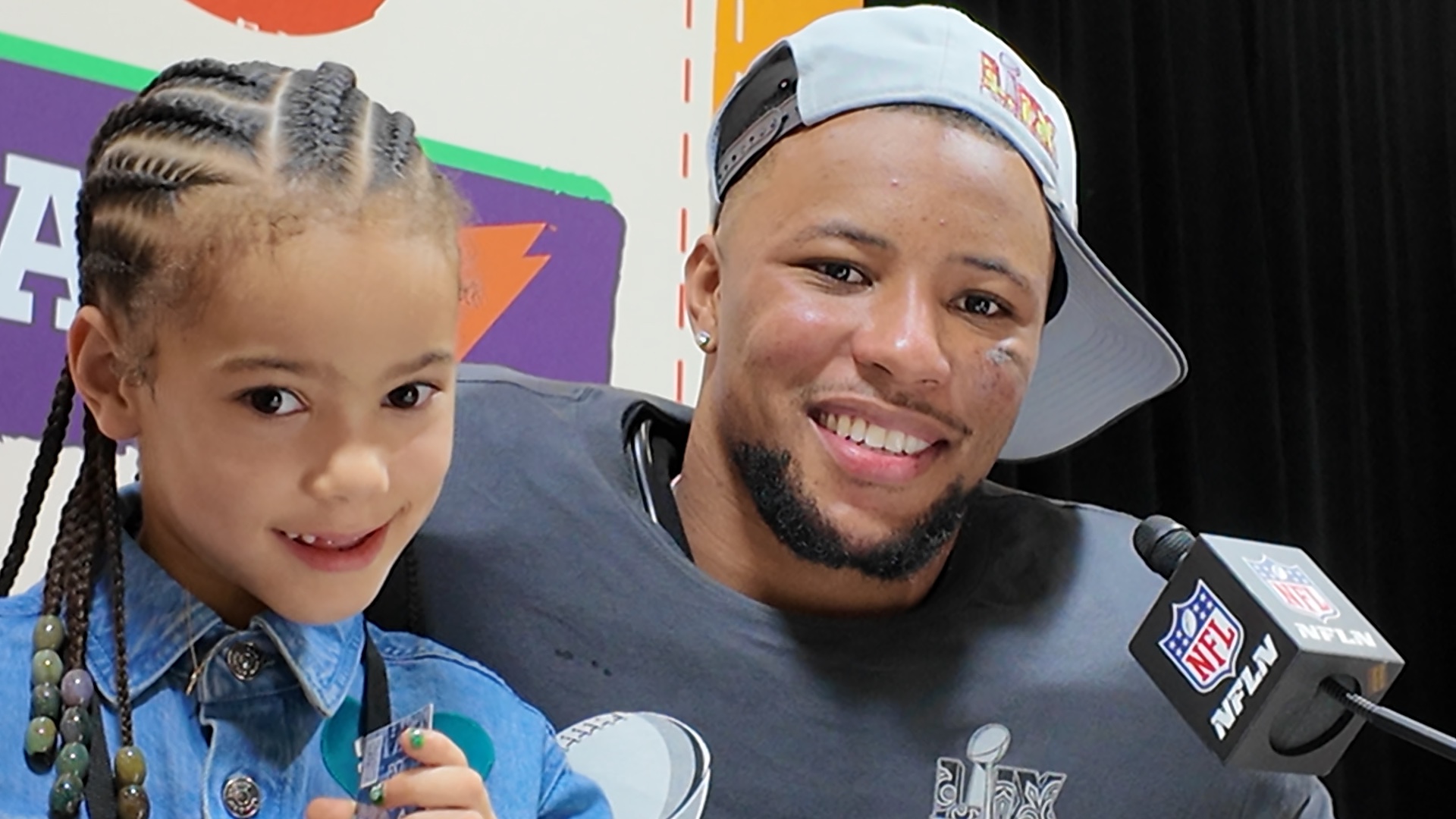 Ein lächelnder Saquon Barkley mit Bart, einer verkehrt herum aufgesetzten Kappe und einem dunklen Hemd mit dem Logo der Philadelphia Eagles posiert mit einem kleinen Mädchen. Das Mädchen mit geflochtenen Haaren und einem blauen Jeanshemd hält einen kleinen Gegenstand in der Hand. Sie sind auf einer Pressekonferenz mit einem farbenfrohen Hintergrund und einem NFL-Mikrofon. Diese Beschreibung wurde mit der FootballR KI automatisch generiert.