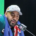 Ein Footballspieler bei einer Pressekonferenz, gekleidet in ein grünes Philadelphia Eagles-Trikot und eine weiße Kappe. Sein Bart blitzt hervor, während er in eine Flagge gehüllt ist. Vor ihm steht ein Mikrofon mit NFL-Logo bereit. Der schwarze Hintergrund wird durch einen grünen Abschnitt mit leuchtend gelbem Super Bowl-Text akzentuiert. Diese Beschreibung wurde mit der FootballR KI automatisch generiert.