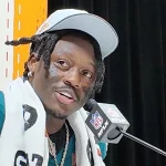 Bei der Pressekonferenz sitzt ein Footballspieler der Philadelphia Eagles in seinem grünen Trikot mit der „24“ selbstbewusst mit einem Handtuch über den Schultern. Mit schwarzer Gesichtsbemalung unter den Augen und einer weißen Kappe spricht er vor einem verschwommenen orange-weißen Hintergrund in ein Mikrofon mit NFL-Logo und hofft auf den Sieg im Super Bowl. Diese Beschreibung wurde mit der FootballR KI automatisch generiert.