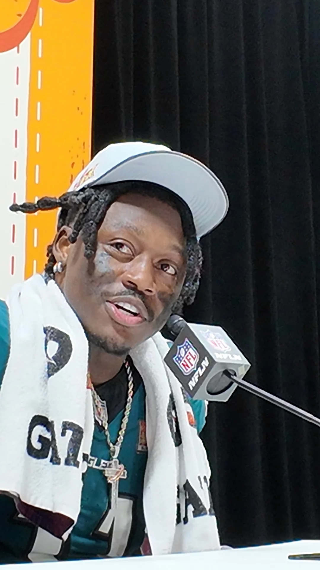 Bei der Pressekonferenz sitzt ein Footballspieler der Philadelphia Eagles in seinem grünen Trikot mit der „24“ selbstbewusst mit einem Handtuch über den Schultern. Mit schwarzer Gesichtsbemalung unter den Augen und einer weißen Kappe spricht er vor einem verschwommenen orange-weißen Hintergrund in ein Mikrofon mit NFL-Logo und hofft auf den Sieg im Super Bowl. Diese Beschreibung wurde mit der FootballR KI automatisch generiert.