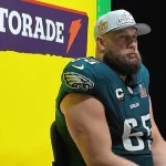 Ein Footballspieler in der Uniform der Philadelphia Eagles sitzt selbstbewusst bei einer Pressekonferenz. Er trägt eine weiße Kappe und die Trikotnummer 65 und blickt den Medien entgegen, während die Vorfreude auf den Super Bowl steigt. Das ikonische leuchtend gelbe Gatorade-Schild steht prominent hinter ihm und umrahmt die Szene. Diese Beschreibung wurde mit der FootballR KI automatisch generiert.
