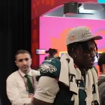 Ein Footballspieler in der Uniform der Philadelphia Eagles steht im Gebäude. Er trägt stolz eine „Super Bowl Champions“-Kappe und ein Handtuch um den Hals. Menschen versammeln sich um ihn, darunter ein Mann im Anzug und jemand mit einem strahlenden Lächeln, der eine Kamera hält. Im Hintergrund feiern bunte Banner den Sieg. Diese Beschreibung wurde mit der FootballR KI automatisch generiert.