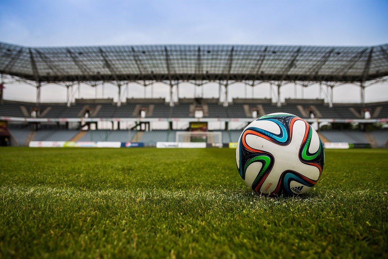 Eine Nahaufnahme eines bunten Fußballs mit weißer Grundfläche und blauen, orangen und grünen Mustern liegt auf einem gepflegten grünen Feld. Im Hintergrund steht ein leeres Stadion unter einem klaren Himmel und weckt Erinnerungen an Topstürmer-Momente der österreichischen Fußball-Ikone Toni Polster. Diese Beschreibung wurde mit der FootballR KI automatisch generiert.