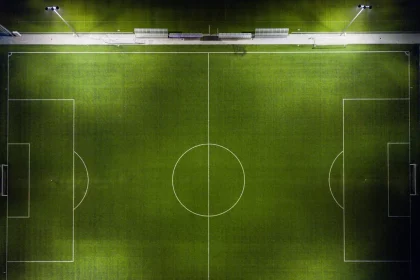 Luftaufnahme eines gut beleuchteten, leeren Fußballfelds bei Nacht. Das grüne Spielfeld zeigt klare weiße Begrenzungslinien, einen Mittelkreis und Strafräume. Vier Flutlichter beleuchten das Feld und werfen Schatten. Außerhalb des Felds sind Bänke und Wege sichtbar. Es sind keine Menschen anwesend. Diese Beschreibung wurde mit der FootballR KI automatisch generiert.