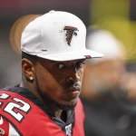 An der Seitenlinie steht ein Footballspieler in einem rot-schwarzen Trikot mit der Nummer 22, Keanu Neal. Er trägt eine weiße Kappe mit dem Logo seines Teams. Der Hintergrund verschwimmt zu undeutlichen Formen und Farben, während sein Gesichtsausdruck ernst bleibt. Diese Beschreibung wurde mit der FootballR KI automatisch generiert.