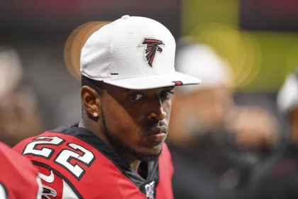 An der Seitenlinie steht ein Footballspieler in einem rot-schwarzen Trikot mit der Nummer 22, Keanu Neal. Er trägt eine weiße Kappe mit dem Logo seines Teams. Der Hintergrund verschwimmt zu undeutlichen Formen und Farben, während sein Gesichtsausdruck ernst bleibt. Diese Beschreibung wurde mit der FootballR KI automatisch generiert.