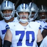 In einem Tunnel stehen die Dallas Cowboys vereint in ihren Uniformen. In der Mitte trägt Zack Martin einen Helm mit Gesichtsschutz und zeigt stolz die Nummer 70 auf seinem Trikot. Hinter ihm bereiten sich seine Mitspieler darauf vor, das Spielfeld zu stürmen. Diese Beschreibung wurde mit der FootballR KI automatisch generiert.