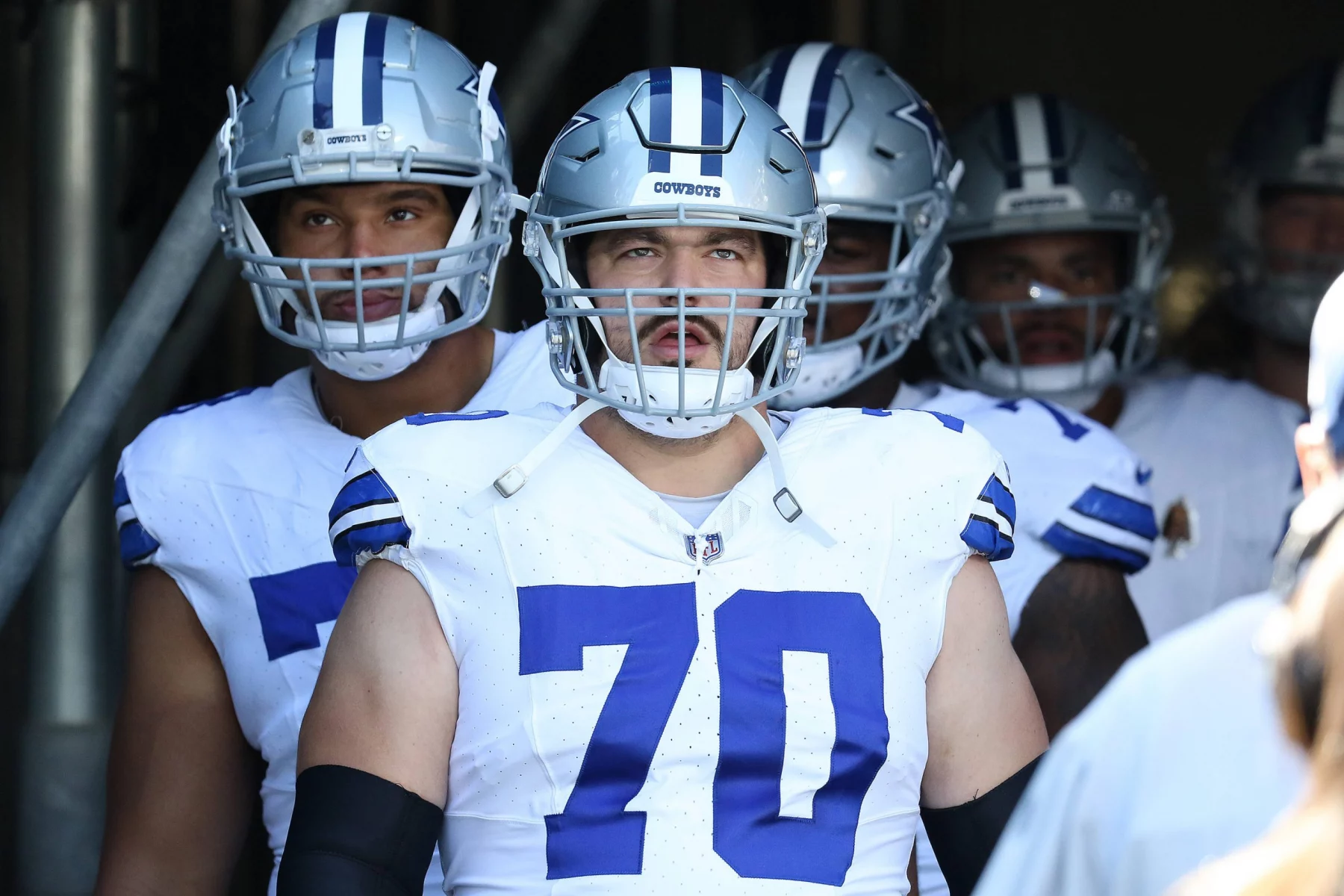 In einem Tunnel stehen die Dallas Cowboys vereint in ihren Uniformen. In der Mitte trägt Zack Martin einen Helm mit Gesichtsschutz und zeigt stolz die Nummer 70 auf seinem Trikot. Hinter ihm bereiten sich seine Mitspieler darauf vor, das Spielfeld zu stürmen. Diese Beschreibung wurde mit der FootballR KI automatisch generiert.
