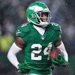 In einem grünen Trikot mit der Nummer 24 und einem Adlerlogo rennt James Bradberry mit dem Football in der Hand über das nasse Feld. Sein grüner Helm und seine weißen Handschuhe fallen im Regen auf und sorgen für eine dynamische Atmosphäre. Ein weiterer Spieler in ähnlicher Kleidung ist links teilweise zu sehen. Diese Beschreibung wurde mit der FootballR KI automatisch generiert.