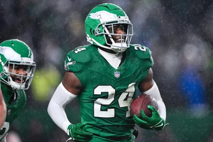 In einem grünen Trikot mit der Nummer 24 und einem Adlerlogo rennt James Bradberry mit dem Football in der Hand über das nasse Feld. Sein grüner Helm und seine weißen Handschuhe fallen im Regen auf und sorgen für eine dynamische Atmosphäre. Ein weiterer Spieler in ähnlicher Kleidung ist links teilweise zu sehen. Diese Beschreibung wurde mit der FootballR KI automatisch generiert.