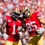 Zwei Footballspieler in den roten Trikots der San Francisco 49ers umarmen sich auf dem Spielfeld. George Kittle, der einen Football hält, beugt sich zu seinem Teamkollegen Deebo Samuel, während sie jubeln. Im Hintergrund ist eine verschwommene Menschenmenge in einem Stadion zu sehen, die die Spannung des Spiels einfängt. Diese Beschreibung wurde mit der FootballR KI automatisch generiert.