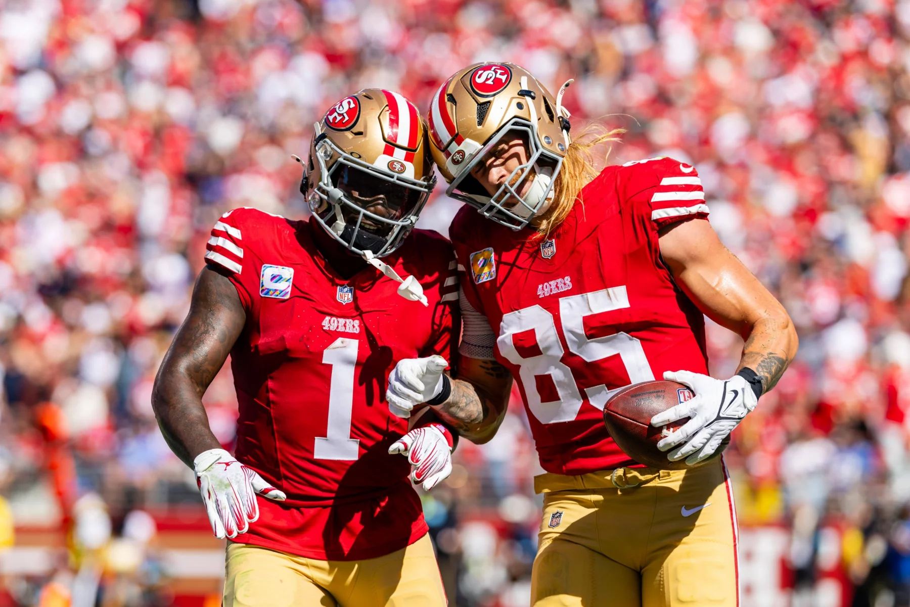 Zwei Footballspieler in den roten Trikots der San Francisco 49ers umarmen sich auf dem Spielfeld. George Kittle, der einen Football hält, beugt sich zu seinem Teamkollegen Deebo Samuel, während sie jubeln. Im Hintergrund ist eine verschwommene Menschenmenge in einem Stadion zu sehen, die die Spannung des Spiels einfängt. Diese Beschreibung wurde mit der FootballR KI automatisch generiert.