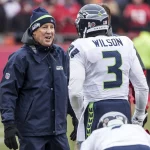 Ein Footballtrainer in marineblauer Jacke und Mütze spricht mit Russell Wilson, Nummer 3, in einer weißen Uniform der Seattle Seahawks. Sie stehen an einem bewölkten Tag auf einem Spielfeld mit verschwommenen Zuschauern im Hintergrund. Der Trainer wirkt lebhaft, während er mit Wilson spricht. Diese Beschreibung wurde mit der FootballR KI automatisch generiert.