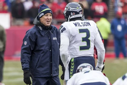 Ein Footballtrainer in marineblauer Jacke und Mütze spricht mit Russell Wilson, Nummer 3, in einer weißen Uniform der Seattle Seahawks. Sie stehen an einem bewölkten Tag auf einem Spielfeld mit verschwommenen Zuschauern im Hintergrund. Der Trainer wirkt lebhaft, während er mit Wilson spricht. Diese Beschreibung wurde mit der FootballR KI automatisch generiert.