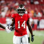 Chris Godwin mit der Nummer 14 in der roten Uniform der Tampa Bay Buccaneers streckt seine rechte Hand zum Händedruck auf dem Spielfeld aus. Chris Godwin trägt einen Helm mit Gesichtsschutz und fingerlose Handschuhe und glänzt in seinem neuen Vertrag, während die verschwommene Menge die Sitze im Stadion füllt. Diese Beschreibung wurde mit der FootballR KI automatisch generiert.