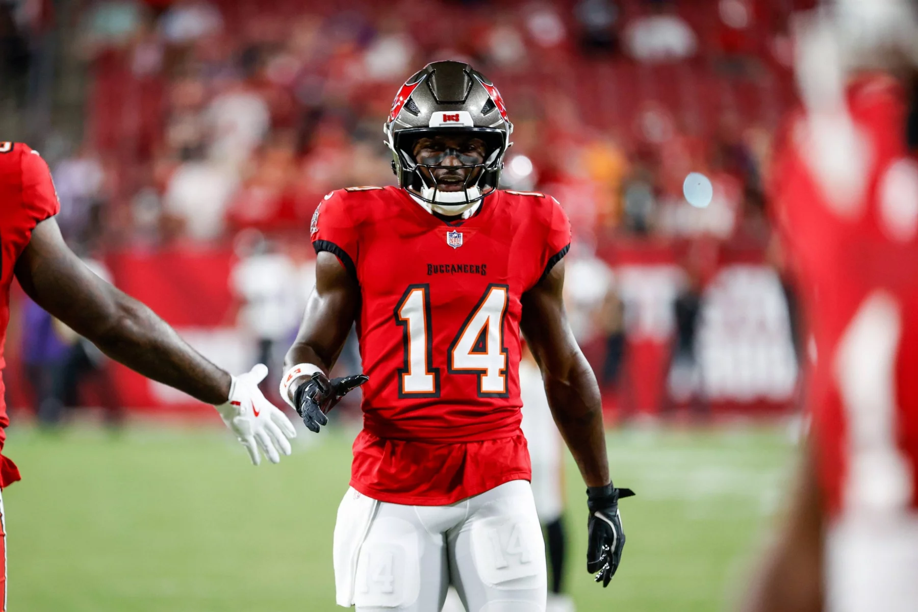 Chris Godwin mit der Nummer 14 in der roten Uniform der Tampa Bay Buccaneers streckt seine rechte Hand zum Händedruck auf dem Spielfeld aus. Chris Godwin trägt einen Helm mit Gesichtsschutz und fingerlose Handschuhe und glänzt in seinem neuen Vertrag, während die verschwommene Menge die Sitze im Stadion füllt. Diese Beschreibung wurde mit der FootballR KI automatisch generiert.
