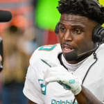 Tyreek Hill in weißer Miami Dolphins-Uniform, mit Helm und Mikrofon-Headset, spricht lebhaft in die Kamera. Seine behandschuhte Hand gestikuliert beim Sprechen. Ist das Tyreek Hill? Im Hintergrund sind verschwommene Stadionsitze und Beschilderungen zu sehen, was auf eine Interview-Situation nach dem Spiel hindeutet. Diese Beschreibung wurde mit der FootballR KI automatisch generiert.