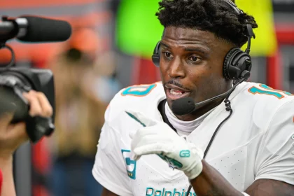 Tyreek Hill in weißer Miami Dolphins-Uniform, mit Helm und Mikrofon-Headset, spricht lebhaft in die Kamera. Seine behandschuhte Hand gestikuliert beim Sprechen. Ist das Tyreek Hill? Im Hintergrund sind verschwommene Stadionsitze und Beschilderungen zu sehen, was auf eine Interview-Situation nach dem Spiel hindeutet. Diese Beschreibung wurde mit der FootballR KI automatisch generiert.