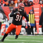 Trey Hendrickson trägt die Nummer 91 und rennt in seiner schwarz-orangen Uniform der Cincinnati Bengals über das grüne Feld. Mit seinem gestreiften Helm verkörpert er Agilität und Geschicklichkeit zugleich. Hinter ihm ist eine geschäftige Seitenlinie aus Offiziellen und Mitarbeitern durch leuchtend orangefarbene Markierungen hervorgehoben, während die Menge zu einem verschwommenen Bild verschwimmt. Diese Beschreibung wurde mit der FootballR KI automatisch generiert.