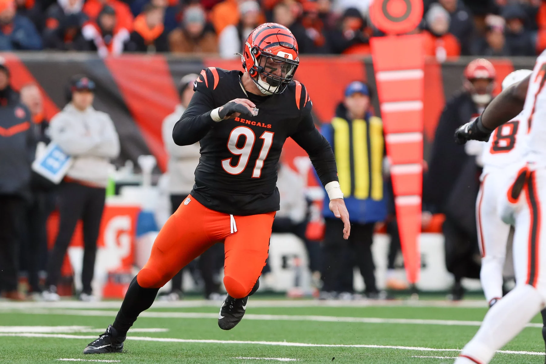Trey Hendrickson trägt die Nummer 91 und rennt in seiner schwarz-orangen Uniform der Cincinnati Bengals über das grüne Feld. Mit seinem gestreiften Helm verkörpert er Agilität und Geschicklichkeit zugleich. Hinter ihm ist eine geschäftige Seitenlinie aus Offiziellen und Mitarbeitern durch leuchtend orangefarbene Markierungen hervorgehoben, während die Menge zu einem verschwommenen Bild verschwimmt. Diese Beschreibung wurde mit der FootballR KI automatisch generiert.