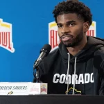 Shedeur Sanders mit kurzen lockigen Haaren und Bart, der einen schwarzen „COLORADO“-Kapuzenpulli trägt, wird bei der Pressekonferenz als Shedeur Sanders identifiziert. Er sitzt hinter einem Mikrofon und einem Namensschild vor einem blauen Hintergrund, der mit Valero Alamo Bowl-Logos geschmückt ist. Diese Beschreibung wurde mit der FootballR KI automatisch generiert.