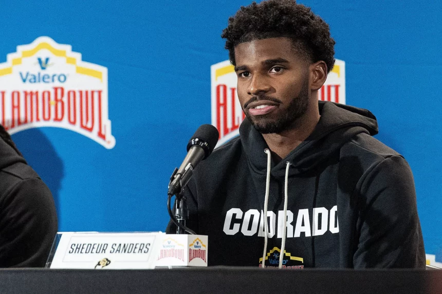 Shedeur Sanders mit kurzen lockigen Haaren und Bart, der einen schwarzen „COLORADO“-Kapuzenpulli trägt, wird bei der Pressekonferenz als Shedeur Sanders identifiziert. Er sitzt hinter einem Mikrofon und einem Namensschild vor einem blauen Hintergrund, der mit Valero Alamo Bowl-Logos geschmückt ist. Diese Beschreibung wurde mit der FootballR KI automatisch generiert.
