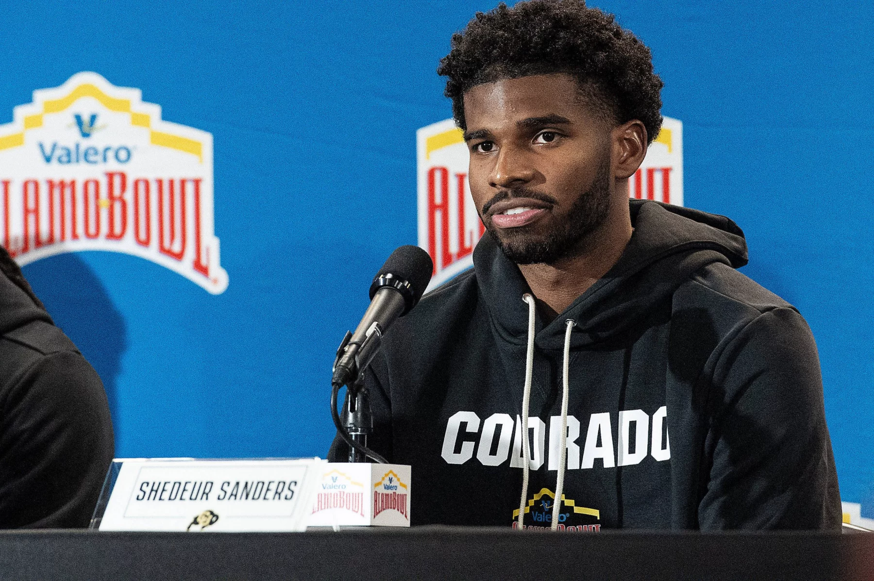 Shedeur Sanders mit kurzen lockigen Haaren und Bart, der einen schwarzen „COLORADO“-Kapuzenpulli trägt, wird bei der Pressekonferenz als Shedeur Sanders identifiziert. Er sitzt hinter einem Mikrofon und einem Namensschild vor einem blauen Hintergrund, der mit Valero Alamo Bowl-Logos geschmückt ist. Diese Beschreibung wurde mit der FootballR KI automatisch generiert.