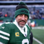 Aaron Rodgers in grün-weißer Uniform, der eine Beanie mit der Aufschrift „Jets“ trägt, steht selbstbewusst auf dem Spielfeld. Mit einem Bart und einem lächelnden Gesicht, das an Aaron Rodgers erinnert, strahlt er vor der verschwommenen Kulisse des Stadions voller eifriger Zuschauer und leuchtender Lichter Energie aus. Diese Beschreibung wurde mit der FootballR KI automatisch generiert.