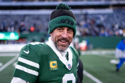 Aaron Rodgers in grün-weißer Uniform, der eine Beanie mit der Aufschrift „Jets“ trägt, steht selbstbewusst auf dem Spielfeld. Mit einem Bart und einem lächelnden Gesicht, das an Aaron Rodgers erinnert, strahlt er vor der verschwommenen Kulisse des Stadions voller eifriger Zuschauer und leuchtender Lichter Energie aus. Diese Beschreibung wurde mit der FootballR KI automatisch generiert.