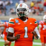 NFL Draft 2025 - Cam Ward in einem orangefarbenen Trikot der University of Miami mit der Nummer 1 und einem weißen Helm, der an Cam Wards Stil erinnert, wärmt sich auf dem Feld auf. Das Trikot weist grüne und weiße Schulterstreifen auf. Andere Spieler und eine verschwommene Menge stehen im Hintergrund, während er auf seine Smartwatch an seinem linken Handgelenk schaut. Diese Beschreibung wurde mit der FootballR KI automatisch generiert.