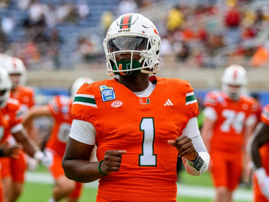 NFL Draft 2025 - Cam Ward in einem orangefarbenen Trikot der University of Miami mit der Nummer 1 und einem weißen Helm, der an Cam Wards Stil erinnert, wärmt sich auf dem Feld auf. Das Trikot weist grüne und weiße Schulterstreifen auf. Andere Spieler und eine verschwommene Menge stehen im Hintergrund, während er auf seine Smartwatch an seinem linken Handgelenk schaut. Diese Beschreibung wurde mit der FootballR KI automatisch generiert.