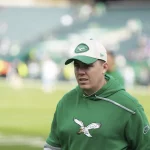 Kellen Moore auf einem Footballfeld trägt einen grünen Kapuzenpullover mit einem Adler-Logo und eine passende Kappe mit einem ähnlichen Logo. Der Hintergrund zeigt ein verschwommenes Stadion mit Reihen leerer Sitze und Menschen, die über das Feld verstreut sind. Die Beleuchtung ist natürlich und deutet auf Tageslicht hin. Diese Beschreibung wurde mit der FootballR KI automatisch generiert.