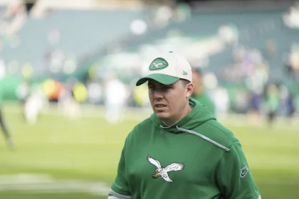 Kellen Moore auf einem Footballfeld trägt einen grünen Kapuzenpullover mit einem Adler-Logo und eine passende Kappe mit einem ähnlichen Logo. Der Hintergrund zeigt ein verschwommenes Stadion mit Reihen leerer Sitze und Menschen, die über das Feld verstreut sind. Die Beleuchtung ist natürlich und deutet auf Tageslicht hin. Diese Beschreibung wurde mit der FootballR KI automatisch generiert.