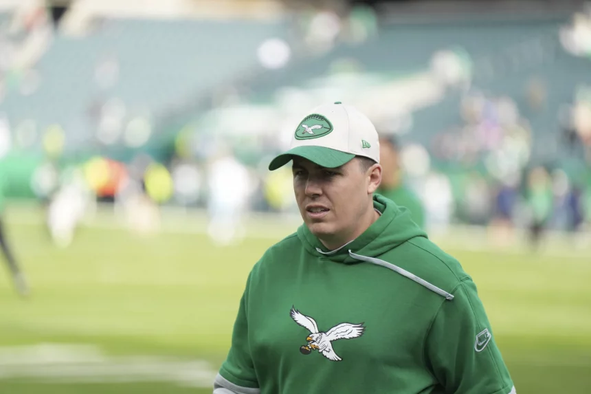 Kellen Moore auf einem Footballfeld trägt einen grünen Kapuzenpullover mit einem Adler-Logo und eine passende Kappe mit einem ähnlichen Logo. Der Hintergrund zeigt ein verschwommenes Stadion mit Reihen leerer Sitze und Menschen, die über das Feld verstreut sind. Die Beleuchtung ist natürlich und deutet auf Tageslicht hin. Diese Beschreibung wurde mit der FootballR KI automatisch generiert.