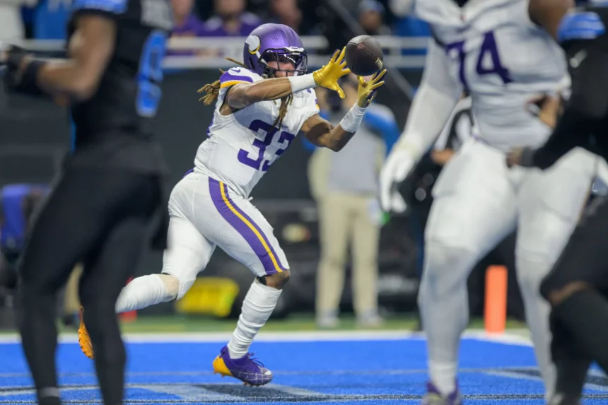Aaron Jones in lila-weißer Uniform, die an den dynamischen Stil von Aaron Jones erinnert, fängt mitten im Spiel einen Pass. Er trägt Helm und Handschuhe und sein Trikot zeigt stolz die Nummer 33. Das Feld hat eine blaue Zone, während Spieler in schwarzen Uniformen im Hintergrund Strategien entwickeln. Diese Beschreibung wurde mit der FootballR KI automatisch generiert.